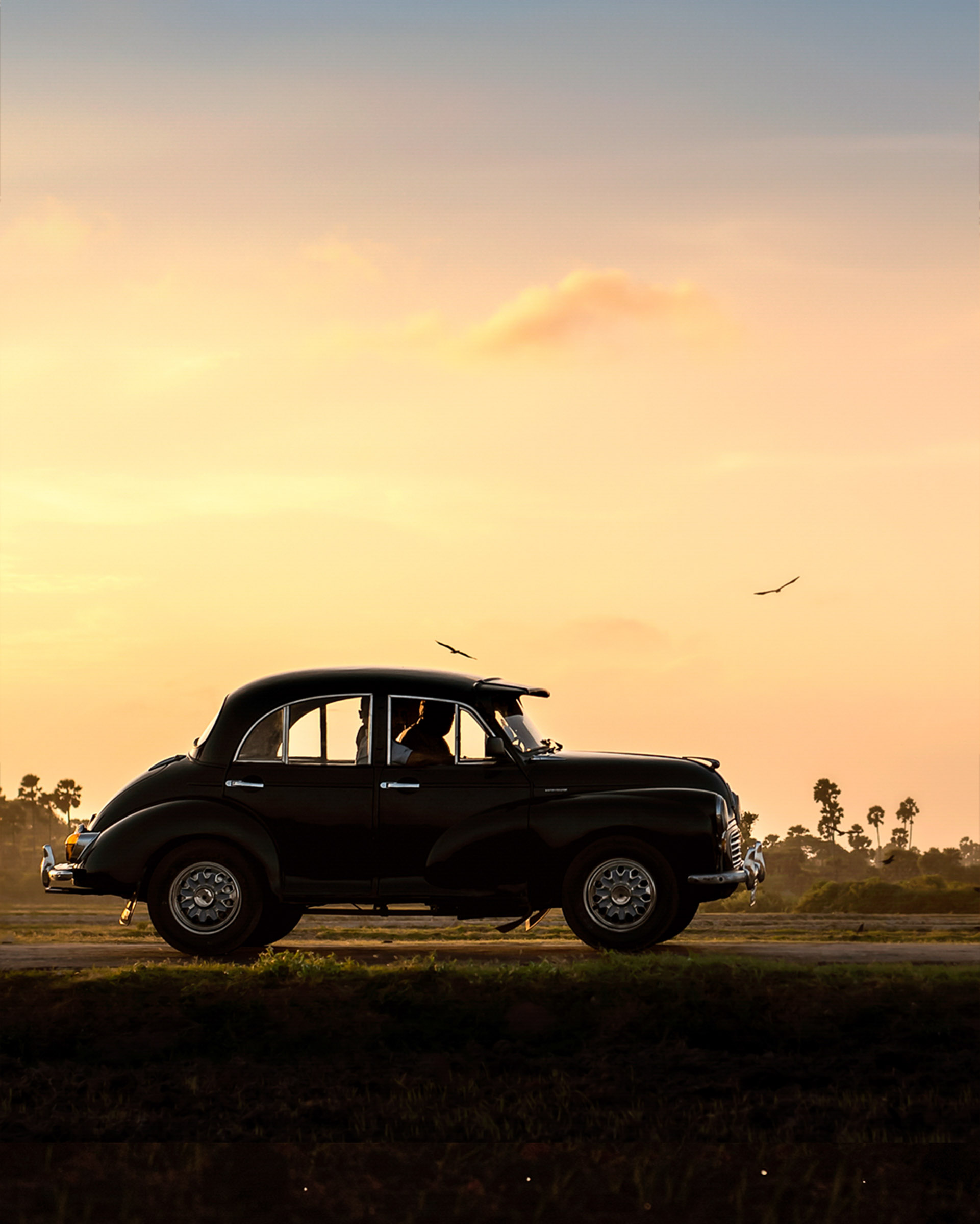 A vintage Morris Minor car driving along a scenic road in Jaffna, Sri Lanka, with lush greenery and traditional architecture in the background.
