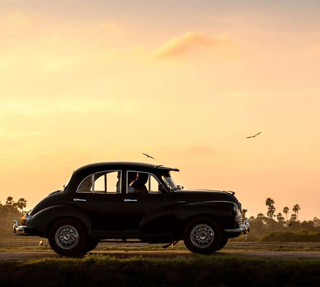 A vintage Morris Minor car driving along a scenic road in Jaffna, Sri Lanka, with lush greenery and traditional architecture in the background.