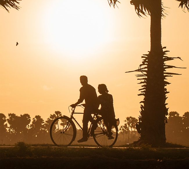 Local Jaffna residents riding bicycles along a road, surrounded by vibrant greenery and traditional Sri Lankan architecture, showcasing the community's everyday life.